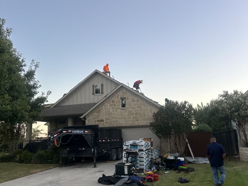 Job site staged for a roof replacement, with a dump trailer and shingle bundles on the driveway.